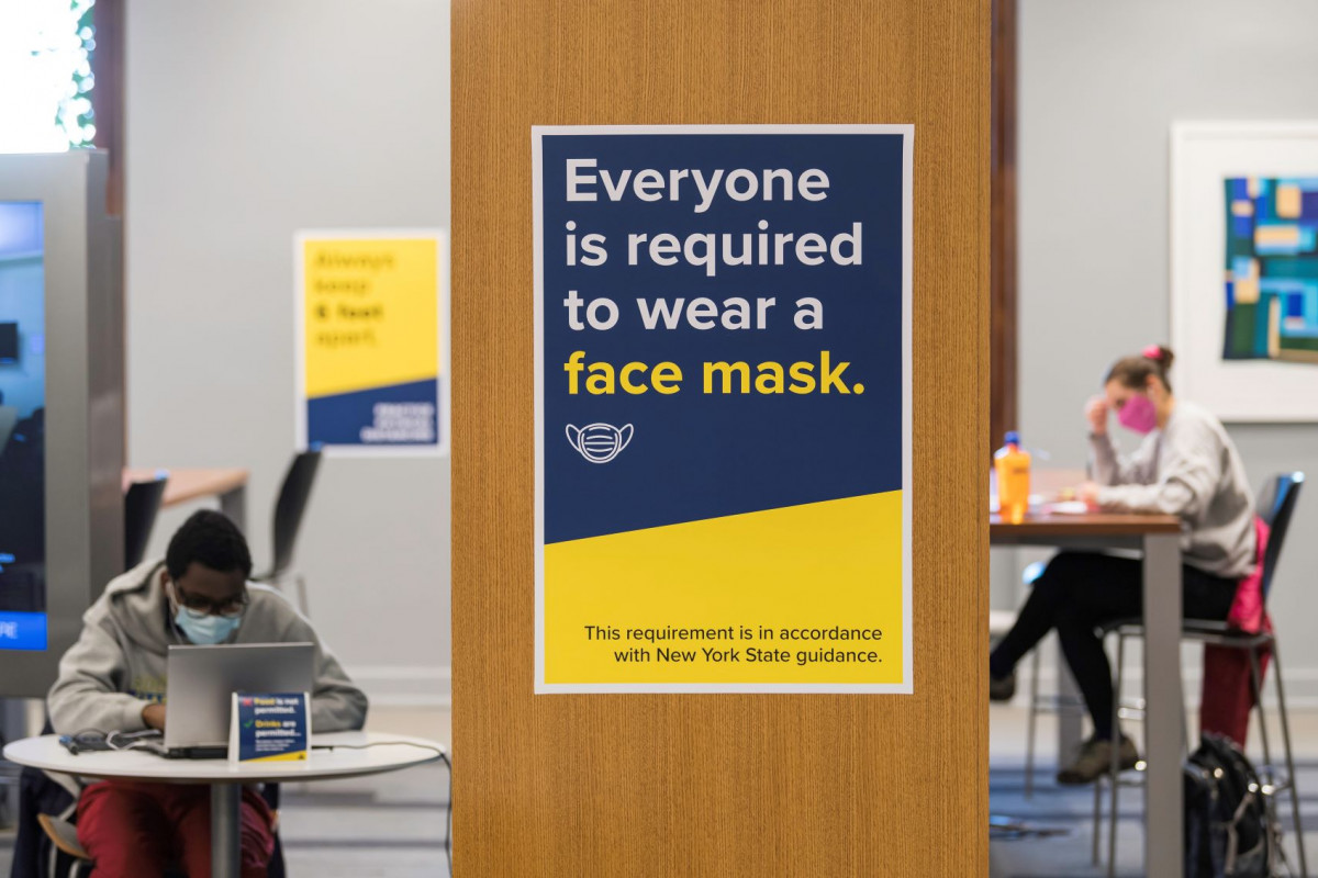 Foreground: a sign saying everyone must wear a mask. Background: Students wearing face masks studying in Evans Lam Square 