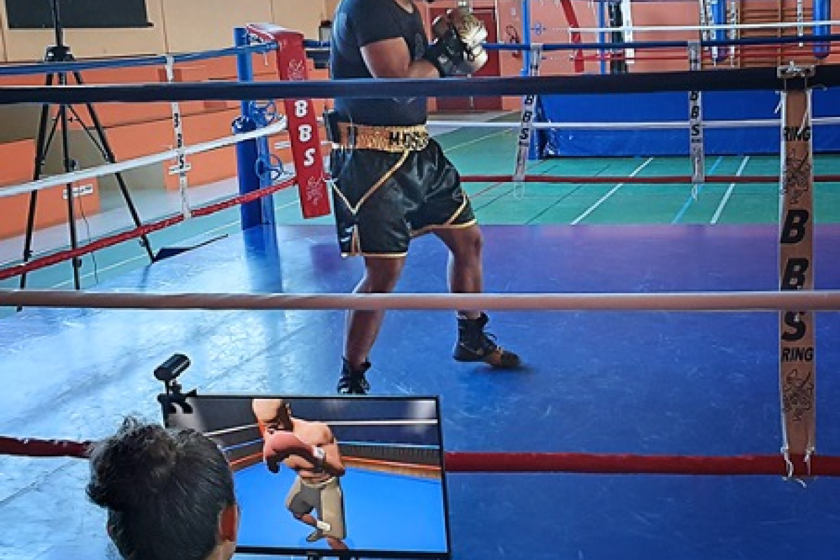 A person wearing a VR headset and boxing gloves trains in a boxing ring while someone monitors their progress on a screen nearby.