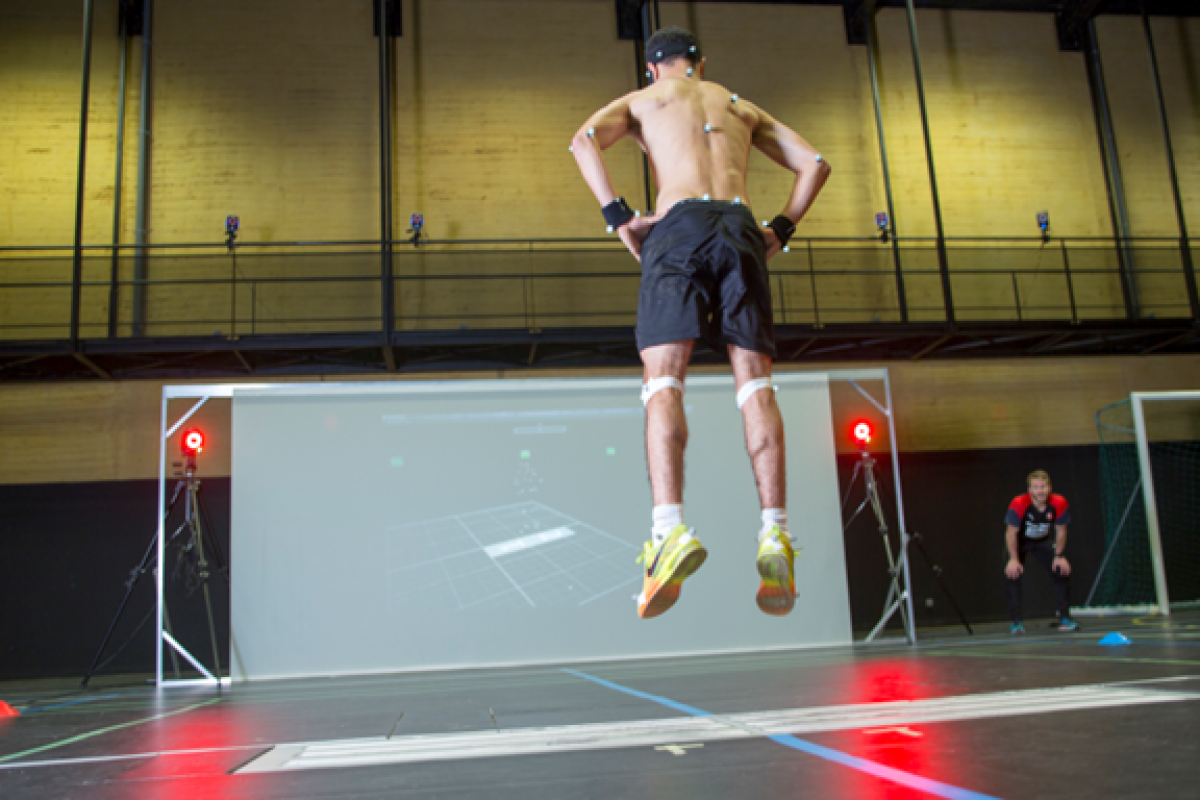A shirtless athlete wearing motion-tracking sensors jumps in front of a large screen displaying his movements, while a coach observes from the side in an indoor sports facility.