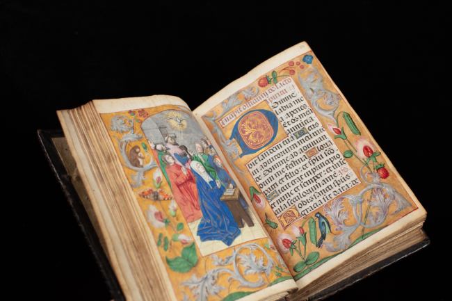 An illuminated manuscript open to two pages, featuring elaborate hand-painted illustrations and calligraphy. The left page shows a colorful religious scene with figures in medieval clothing, surrounded by a gold floral border. The right page contains Latin text in blackletter script with ornate capital letters and a decorated border of flowers, vines, and small birds. The book is displayed against a black background.