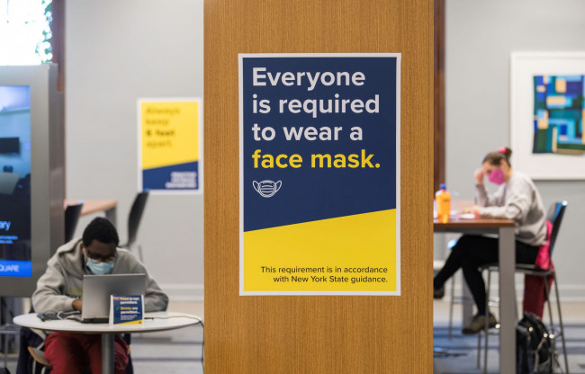 Foreground: a sign saying everyone must wear a mask. Background: Students wearing face masks studying in Evans Lam Square