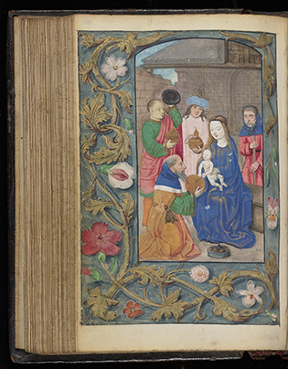 An illuminated manuscript showing a richly colored illustration of the Adoration of the Magi. The Virgin Mary, dressed in blue, holds the baby Jesus while the three Magi present their gifts, kneeling in reverence. Joseph stands behind Mary. The scene is framed with an ornate floral border featuring gold vines, pink and white flowers, and green leaves. The book is slightly open, revealing the thickness of its aged pages